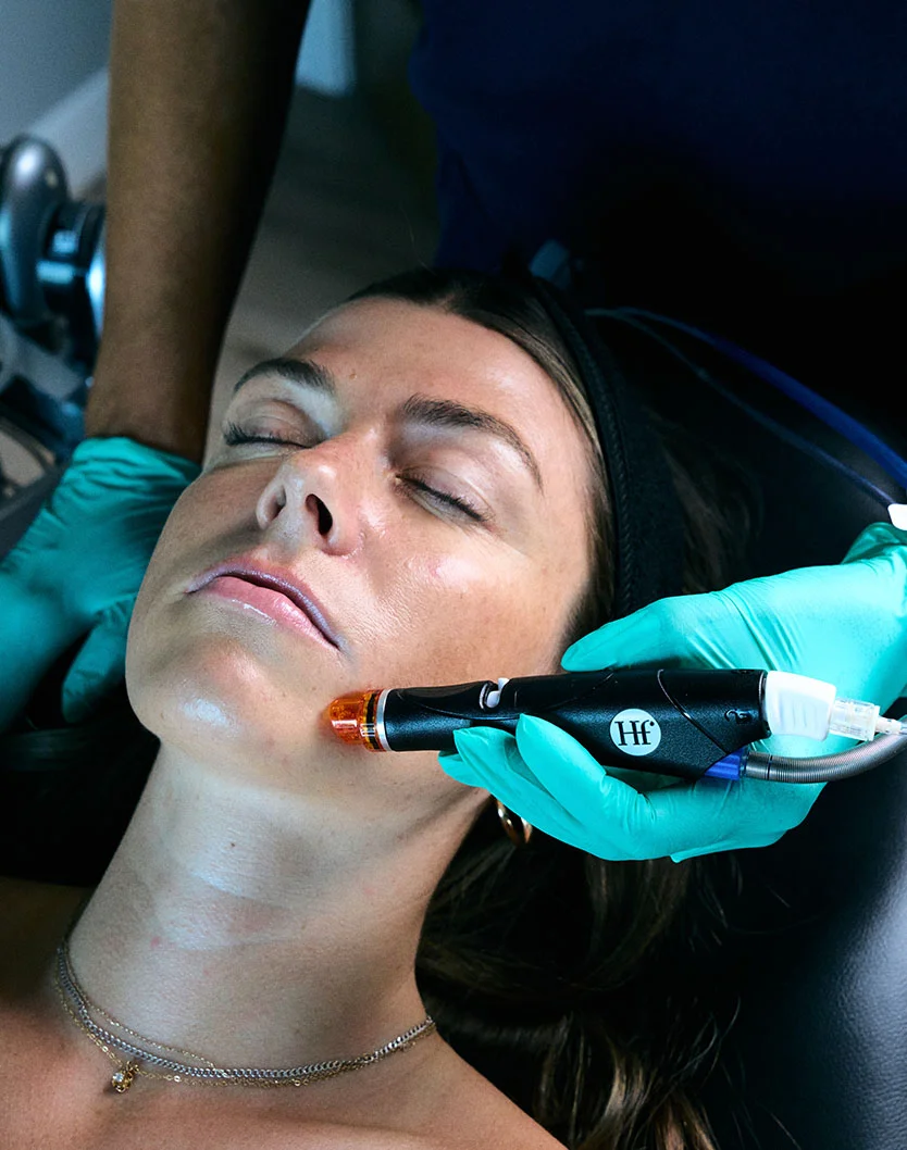 A light-skinned woman with dark hair lies on a treatment table with eyes closed, wearing a dark headband. A medical professional wearing teal gloves holds a dark device with an orange component near the woman’s jawline, performing a facial procedure. The woman wears a multi-chain necklace. Medical equipment and personnel are partially visible in the blurred background. - Hydrafacial in Oconomowoc, WI
