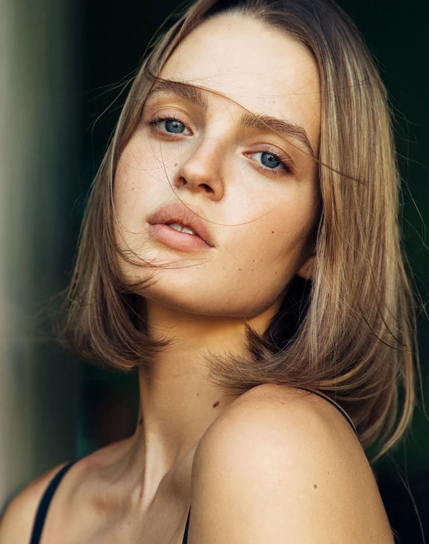 A close-up portrait of a young woman with light brown, shoulder-length hair framing her face. She has smooth, light-toned skin with subtle freckles on her neck and chest. Her neutral expression features soft pink lips and light blue eyes gazing slightly off to the right. She wears a dark sleeveless top against a muted gray-beige background, with natural lighting that highlights her skin’s texture. - Lip Augmentation in Oconomowoc, WI