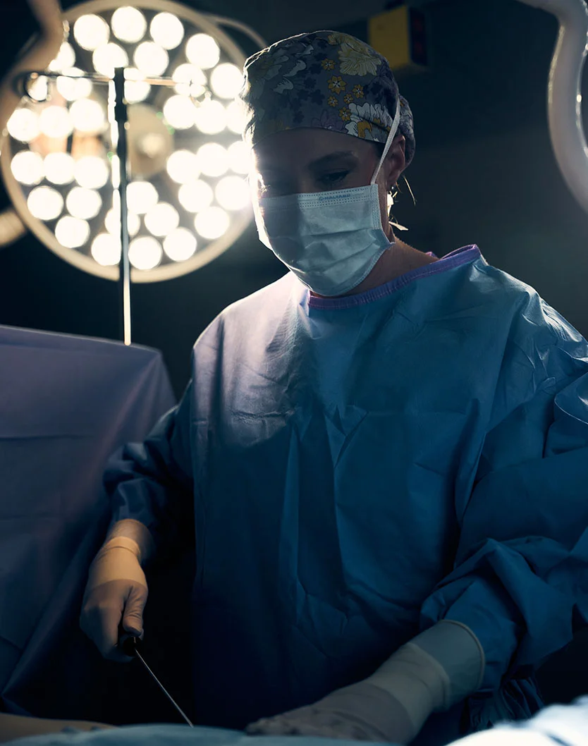 Dr. Parkins, a female surgeon wearing a floral-patterned scrub cap, blue surgical gown, and white surgical mask, is intently focused on a procedure in a dimly lit operating room. She holds a surgical instrument in her gloved hands as a bright overhead light illuminates the surgical field. The rest of the room remains dark, emphasizing the precision and intensity of the moment. - Liposuction in Oconomowoc, WI
