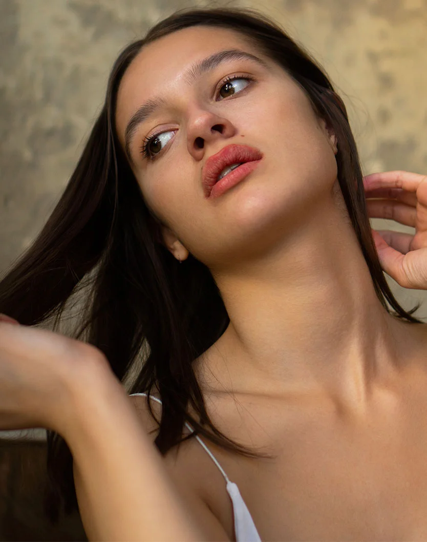 Close-up of a young woman with long, dark brown hair falling around her shoulders. She has smooth, light beige skin with a subtle tan and wears a simple white spaghetti-strap top. Her expression is contemplative, with her gaze directed upward and to the right. Her lips are a soft rosy pink. A warm, soft light illuminates her face and shoulders. The background is a muted, tan-colored wall with a slightly textured surface, and parts of her arms and upper body are visible. - Microneedling in Oconomowoc, WI
