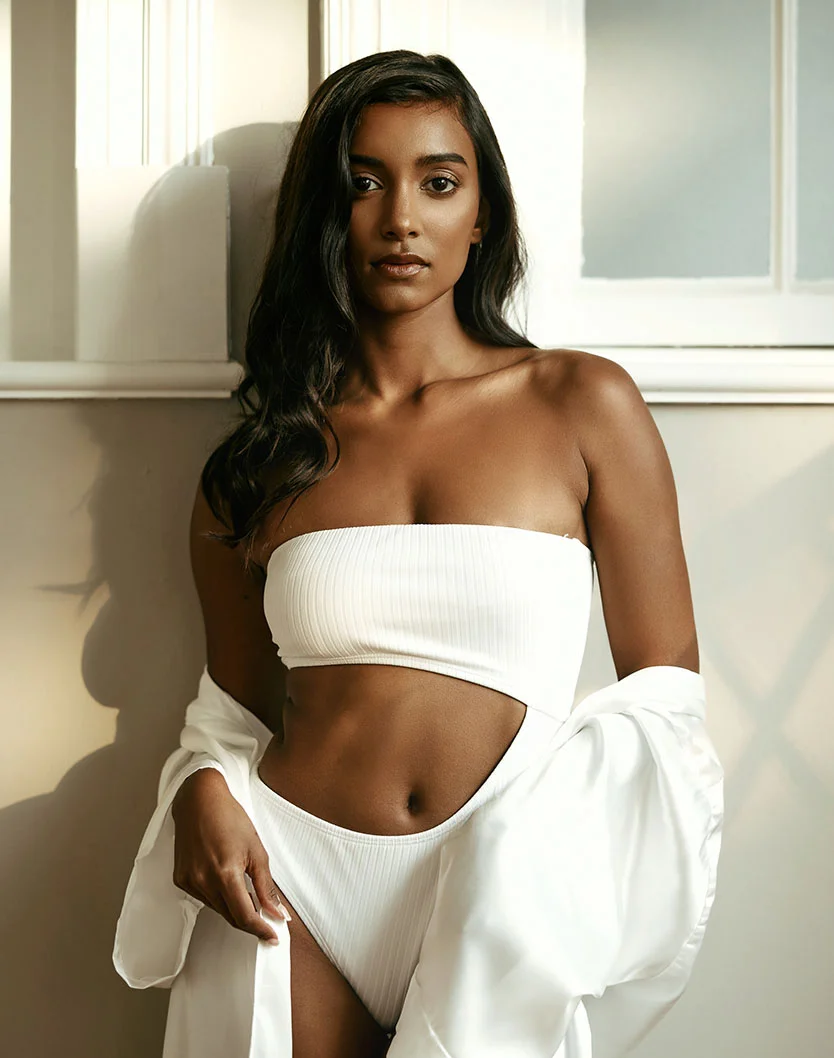 A woman with dark brown skin and long, wavy dark brown hair poses against a light beige wall with white paneled trim. She wears a white, ribbed, strapless top and matching bottoms, resembling a swimsuit or coordinated set, with a subtle texture. A white, oversized silk-like shirt or robe is draped casually over her body. She looks directly at the camera with a neutral expression, exuding relaxed confidence. Soft natural light creates gentle shadows, contributing to a sophisticated and understated aesthetic. - Mommy Makeover in Oconomowoc, WI