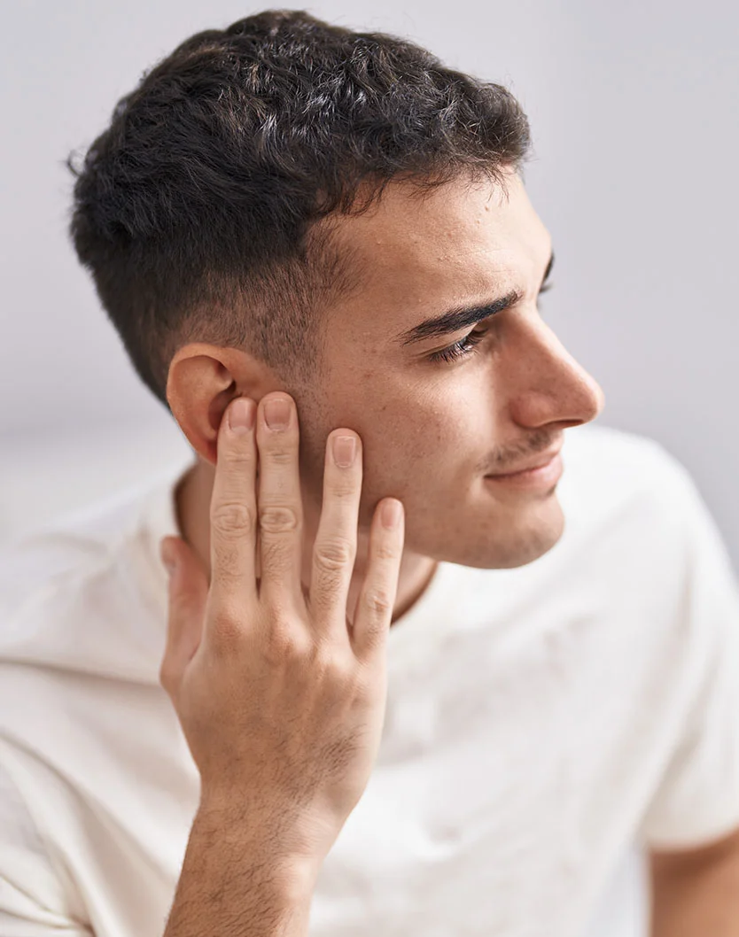 A young man with short, dark curly hair and a light stubble beard is shown in a close-up, looking to his right and smiling slightly. He is wearing a white t-shirt, and his right hand is raised, with fingers spread, touching his earlobe and the side of his face. The background is a soft, light blur. - Otoplasty in Oconomowoc, WI
